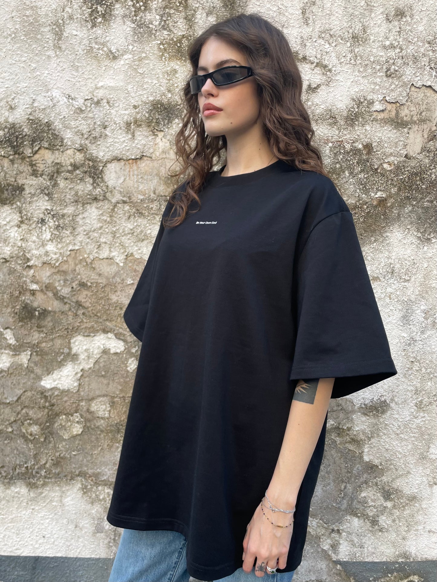 Female model wearing a black heavyweight tee with Be Your Own God front print against a textured wall