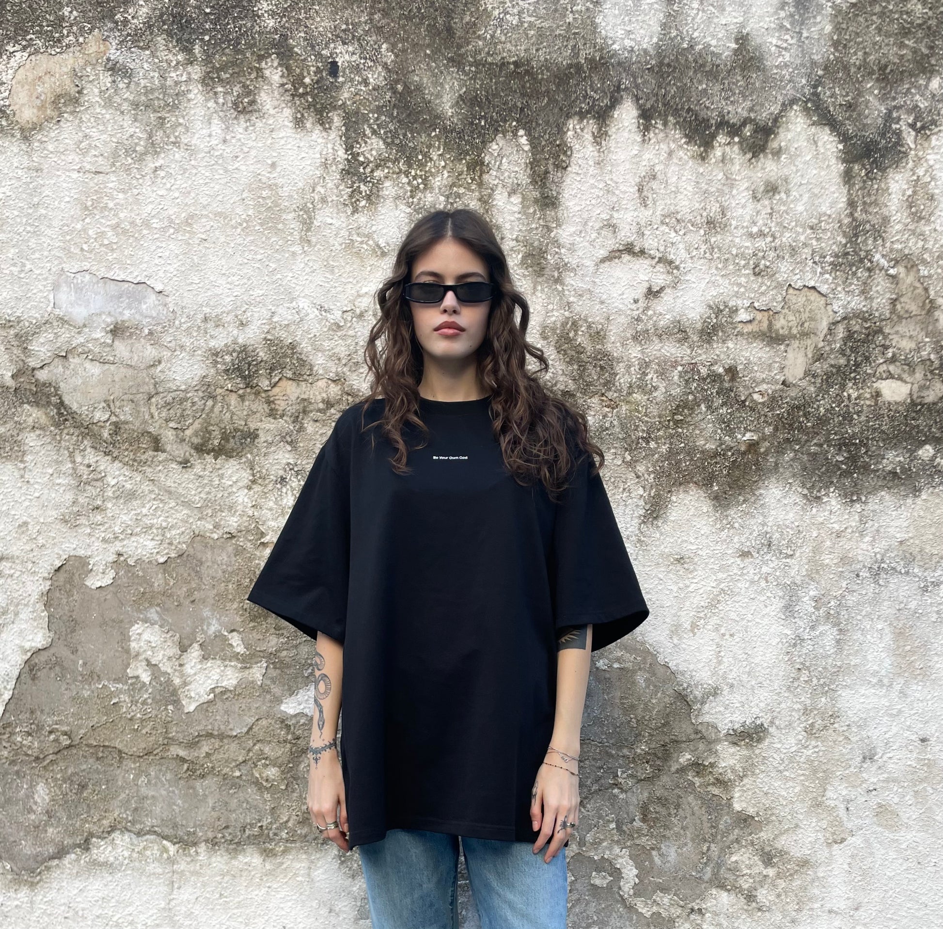 Female model wearing a black heavyweight tee with Be Your Own God front print  against a textured wall