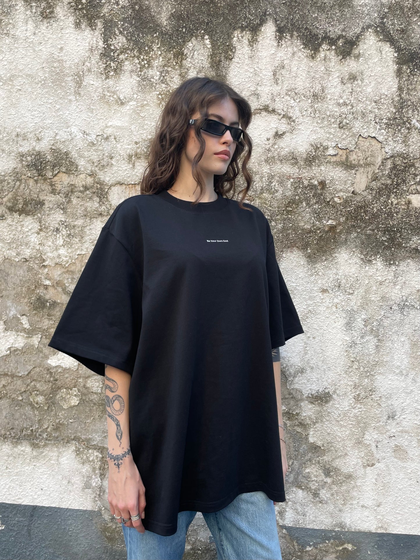 Female model wearing a black heavyweight tee with Be Your Own God front print  against a textured wall