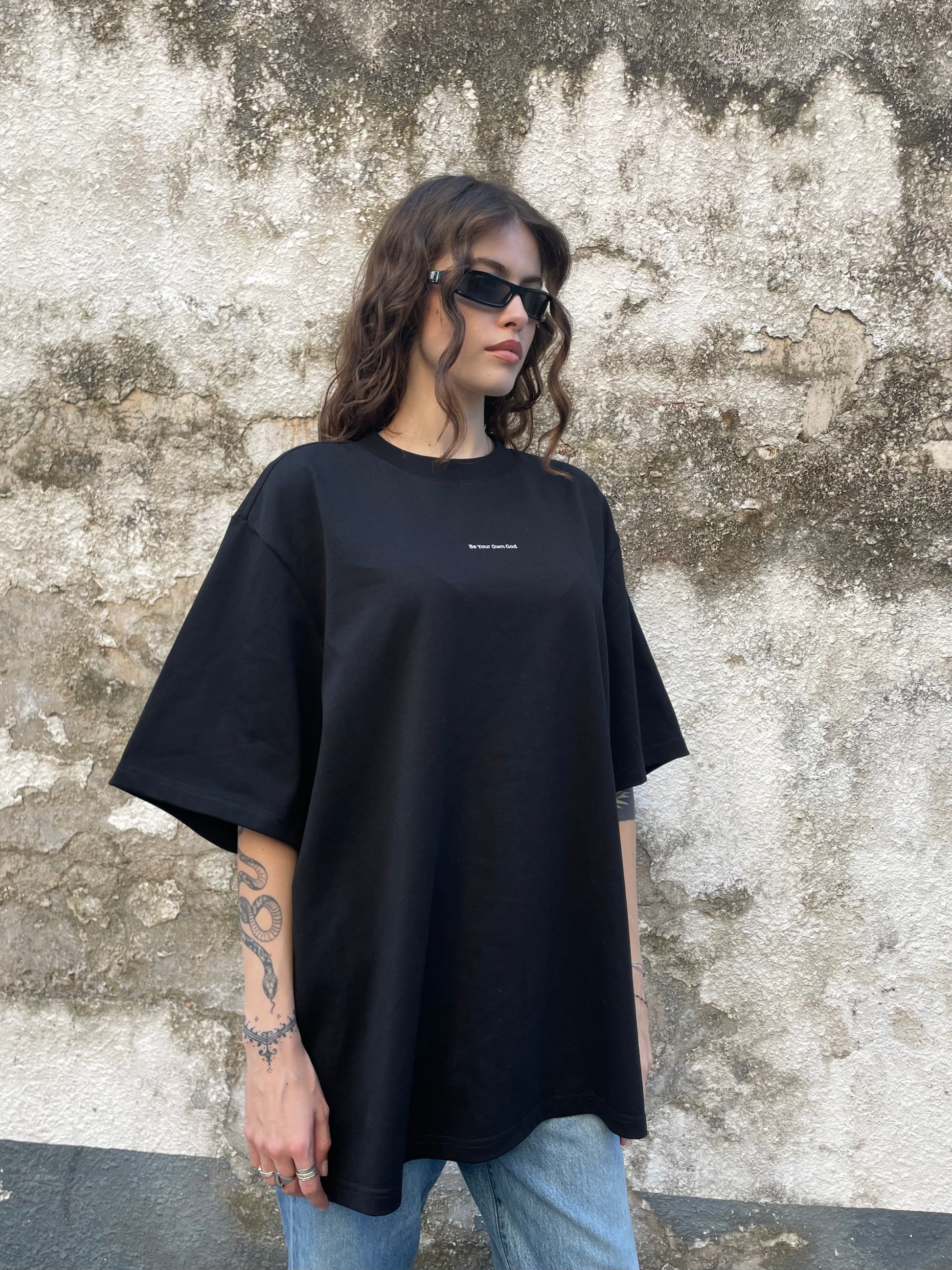 Female model wearing a black heavyweight tee with Be Your Own God front print  against a textured wall