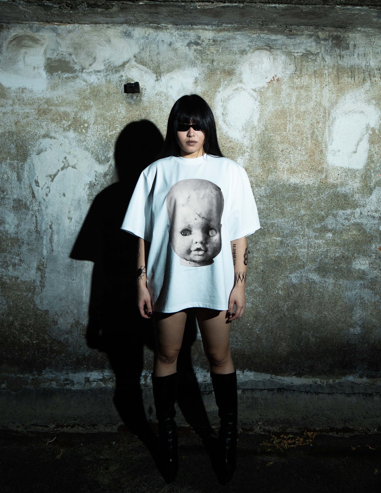 Female Model wearing the BYOG Baby Face white t-shirt in a dark aesthetic setting against a textured wall